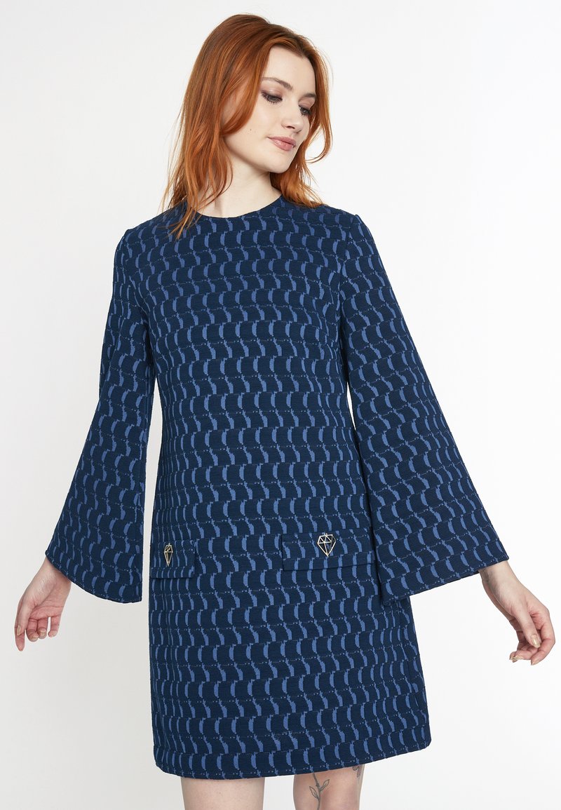 Navyblaues Kleid mit langen Glockenärmeln, strukturiertem Stoff mit geometrischem Muster und zwei Fronttaschen, die mit goldenen Akzenten verziert sind.