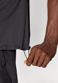 Μαύρο αθλητικό πουκάμισο με κορδόνι στο τελείωμα. Το ύφασμα φαίνεται λείο και ελαφρύ. Κοντινή λήψη χεριού που τραβά το κορδόνι για προσαρμογή.