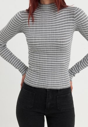 Femme portant un haut à col roulé à manches longues rayé gris et blanc et un jean noir taille haute avec poches, les mains posées sur les hanches.