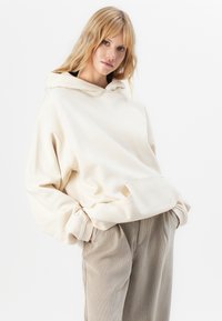 Blonde vrouw draagt een oversized crèmekleurige hoodie met een voorzak en beige corduroy broek, staande tegen een witte achtergrond.