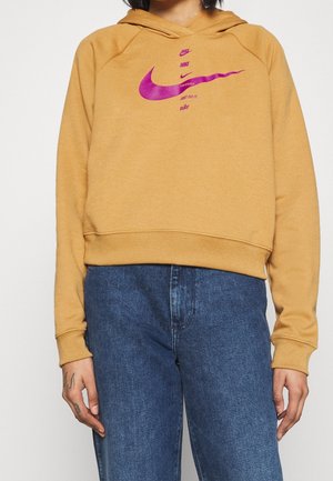 Henkilö pukeutunut sinapinkeltaiseen Nike-huppariin, jossa on useita vaaleanpunaisia Swoosh-logoja ja tekstiä, yhdistettynä sinisiin farkkuihin, jotka näkyvät vyötäröstä alaspäin.