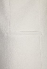 Zblízka vidíme jemnou bílou tkaninu s viditelným švem a malým, horizontálním kapsičkovým detailem. Textura vypadá hřejivě a hladce.