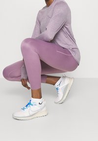 Lilla langærmet atletisk top, matchende lilla leggings og hvide sneakers med blå accenter. Glat stof, tætsiddende design og kort lynlås i siden.
