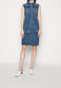 Bezramenné džínové šaty s předním zapínáním na knoflíky, dvěma náprsními kapsami a úzkou sukní. Světle modrá látka s jemnou texturou. Oblečení doplněné bílými otevřenými lodičkami.