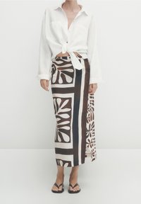 Witte langslavende, gestrikte blouse met twee zakken, gecombineerd met een bruin en crèmewit vloerlange rok met gedurfde, bloemrijke en geometrische patronen.