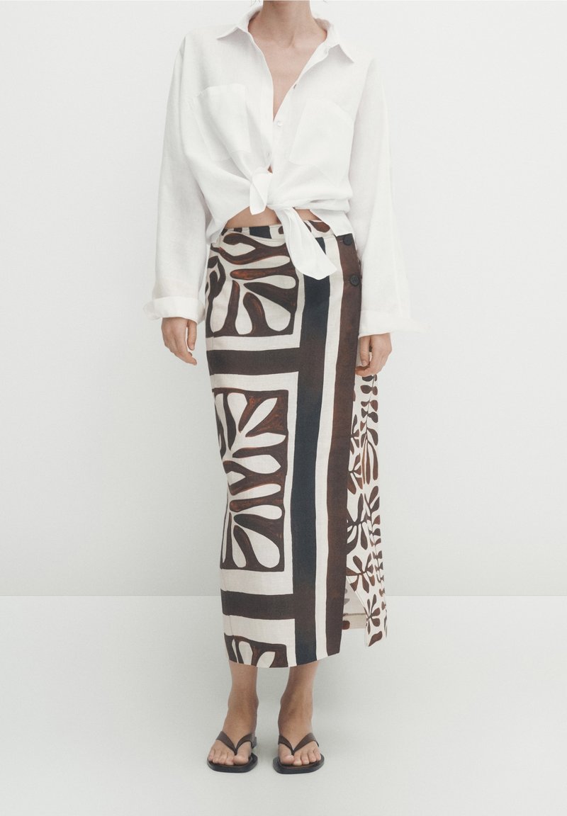 Witte langslavende, gestrikte blouse met twee zakken, gecombineerd met een bruin en crèmewit vloerlange rok met gedurfde, bloemrijke en geometrische patronen.