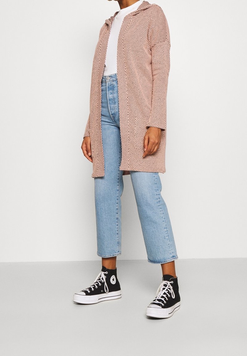 Femme portant un jean court bleu clair, des baskets montantes noires, une chemise blanche et un long cardigan ouvert rose à motifs.
