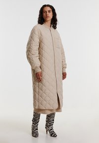 Beige gesteppten langen Mantel mit einem Frontreißverschluss und seitlichem Schlitz. Verfügt über Rippbündchen und eine einzelne Brusttasche. Kombiniert mit Stiefeln im Zebra-Druck.