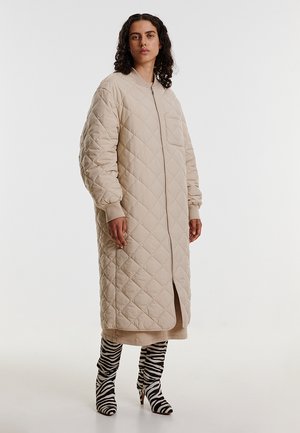 Beige gevoerde lange jas met een voorkant ritssluiting en een zijslit. Voorzien van ribgebreide manchetten en een enkele borstzak. Gecombineerd met laarzen met zebraprint.