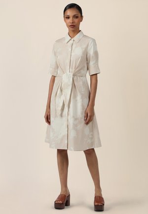 Frau trägt ein beige knielanges Hemdkleid mit Blumenmuster, halblangen Ärmeln und einem gebundenen Taillenband, kombiniert mit braunen Plateausandalen.