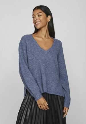 Suéter de punto con escote en V en suave textura azul, con mangas largas y un corte holgado; lo lleva puesto sobre una falda negra con pliegues.