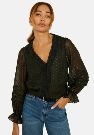 Musta läpinäkyvä paita, jossa on pisteitä, röyhelökaulus ja hihat. Yhdistetty sinisiin farkkuihin ja mustaan vyöhön, jossa on hillittyjä pitsikoristeita.