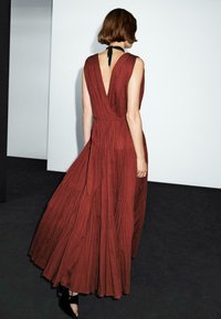 A model wearing a sleeveless, deep-v back, rust-colored maxi dress stands with her back to the camera, showcasing the dress’s flowing fabric.