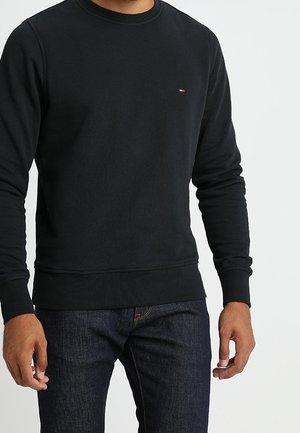 Mężczyzna w prostym czarnym swetrze crewneck z małym logo na piersi, w ciemnoniebieskich dżinsach, stoi na jasnym tle.