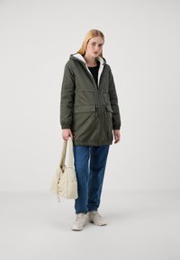 Olivenfarget parka-jakke med hette og faux pelsfôr, med to frontlommer og justerbar snøring i livet. Brukt med blå jeans og en beige veske.