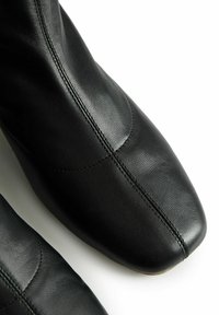 Artimas juodų odinių batų vaizdas, su matoma siūle per centrą ir kreivą siūlę ant balto fono.