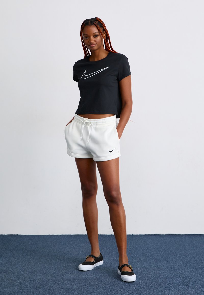 Camiseta negra corta con logo blanco de Nike, pantalones cortos blancos con cordones, y zapatos negros sin cordones con acentos dorados, sobre un fondo liso.