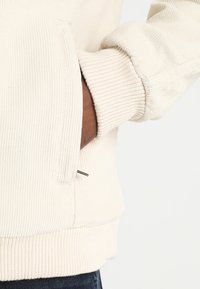 Hand in the side pocket of a light beige corduroy jacket with ribbed cuffs and waistband, showing denim pants below.