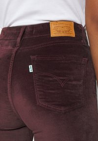 Pantalones de terciopelo burdeos con bolsillos traseros, presentando una etiqueta de cuero marrón deslavada con texto en relieve en la cintura, detalles de costura sutiles.
