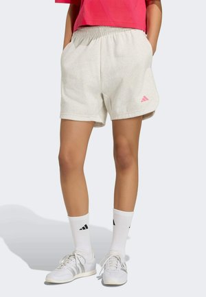Osoba ubrana w jasnobeżowe szorty z różowym logo Adidas, białe skarpetki Adidas, białe trampki i czerwony krótki top, stojąca z rękami w kieszeniach.