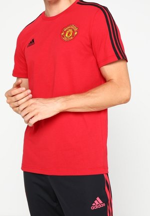T-shirt en coton rouge avec un col rond, rayures noires Adidas sur les épaules et logo de Manchester United sur la poitrine. Manches courtes.