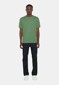 T-shirt verde a maniche corte con collo rotondo e una texture sottile, abbinata a jeans in denim scuri e sneakers bianche.