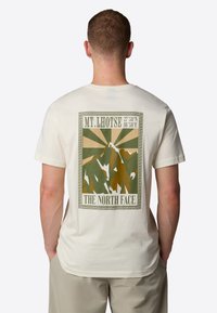 Fuldfarvet t-shirt i cremefarve med et bjergmotiv i jordfarver, med teksten "MT. LHOTSE" og "THE NORTH FACE" ovenfor en dekorativ kant.