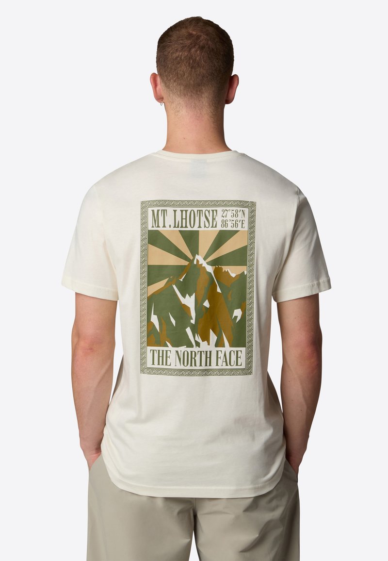 Fuldfarvet t-shirt i cremefarve med et bjergmotiv i jordfarver, med teksten "MT. LHOTSE" og "THE NORTH FACE" ovenfor en dekorativ kant.