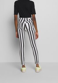 Persona vestida con pantalones de rayas verticales en blanco y negro de cintura alta, camiseta negra de manga corta y zapatillas neón amarillas, de pie de espaldas.