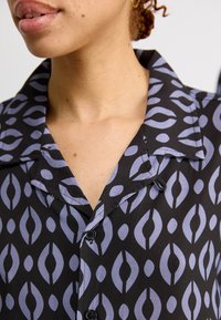 Camiseta negra con un patrón de formas moradas claras superpuestas, con cuello de camisa y detalles de botones. Textura de tejido suave.