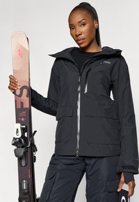 Chaqueta de esquí negra con capucha, cierre con cremallera y varios bolsillos, combinada con esquís que presentan un diseño beige y rosa.