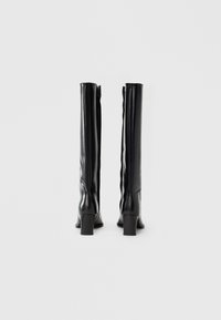 Bottes noires montantes jusqu'en haut de la cuisse en cuir lisse, avec un talon bloc et un détail de fermeture éclair à l'arrière. Design élégant avec une silhouette ajustée.