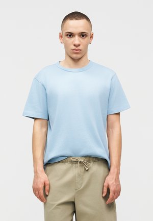 Jeune personne aux cheveux courts portant une chemise bleu clair à manches courtes et un pantalon beige à cordon, debout devant un fond blanc uni.