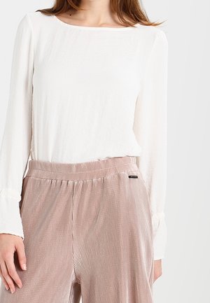Modella che indossa una maglietta bianca a maniche lunghe e pantaloni a palazzo a coste rosa chiaro, in piedi davanti a uno sfondo neutro.