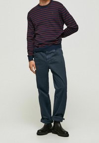 Marinefarbener und roter gestreifter Pullover mit langen Ärmeln, kombiniert mit dunkelblauen, hochgekrempelten Hosen und schwarzen Chelsea-Stiefeln. Glatte Stofftexturen.