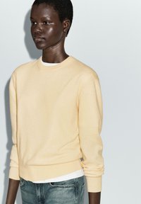 Light yellow crewneck sweater made of soft fabric, paired with a white shirt underneath and fitted blue jeans, showing a smooth texture.