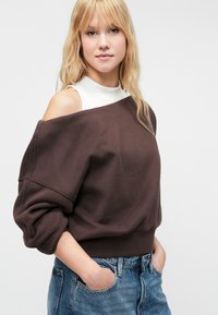 Sudadera marrón con los hombros al descubierto, de mangas largas y con dobladillo acanalado, llevada sobre una blusa blanca de cuello alto. Jeans ajustados azules con bolsillos.