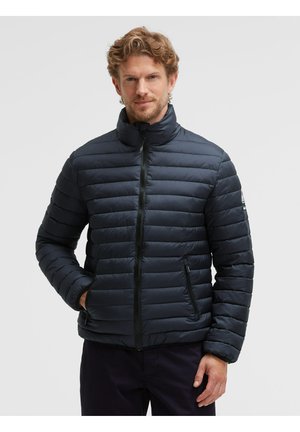 Man with light brown curly hair wearing a zipped black puffer jacket with hands in pockets against a plain light gray background.