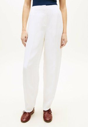 Donna con pantaloni bianchi a gamba larga e vita alta e scarpe marroni con lacci, in piedi contro uno sfondo chiaro e uniforme.