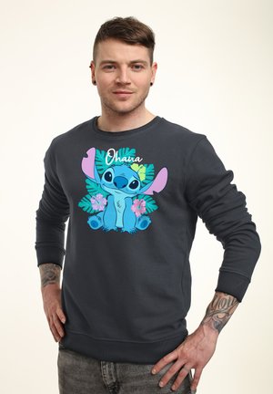 Mężczyzna z tatuażami w czarnej bluzie z kapturem, na której znajduje się niebieska postać z kreskówki Stitch otoczona tropikalnymi liśćmi i kwiatami oraz napisem "Ohana".