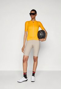 Sárga rövid ujjú kerékpáros mez elöl cipzárral, bézs rövidnadrággal párosítva. A modell fekete sisakot tart, fehér kerékpáros cipőt és mintás zoknit visel.