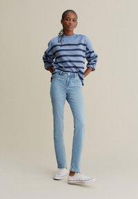 Sweat-shirt à rayures bleu clair avec des rayures bleu marine, associé à un jean bleu clair et des baskets blanches. Le modèle est debout, les mains sur les hanches.