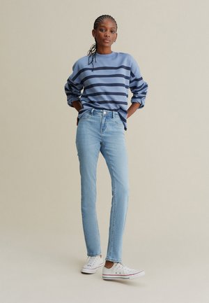 Hanorac cu dungi albastre deschise și dungi bleumarin, asortat cu blugi albastre deschis și adidași albi. Modelul stă cu mâinile pe șolduri.