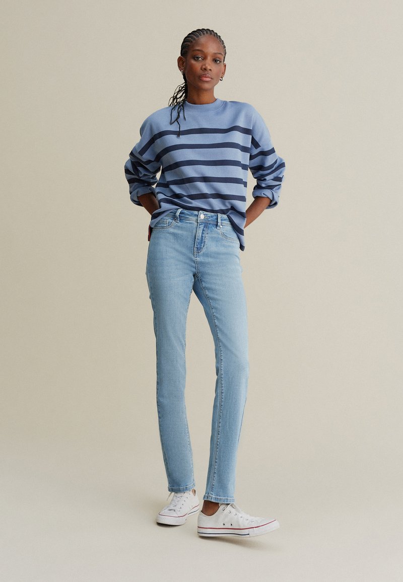 Sweat-shirt à rayures bleu clair avec des rayures bleu marine, associé à un jean bleu clair et des baskets blanches. Le modèle est debout, les mains sur les hanches.