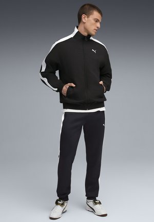 Sort sportsjakke med hvide striber, lynlås foran og elastiske manchetter, parret med mørke bukser med sideløbende striber, båret med hvide sneakers.