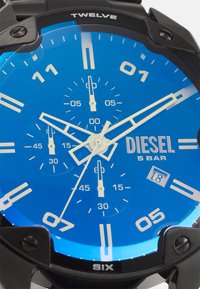 Schwarze Uhr mit großem blauen Zifferblatt, weißen Stundenmarkierungen, Chronographen-Subzifferblättern und einem Datumsdisplay. Wasserdicht bis 5 bar. Besitzt markante Akzente.