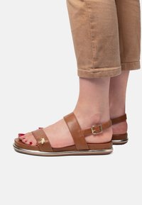 Hnědé kožené sandály se dv�ěma širokými pásky, s detailním sponkovým zapínáním a kovovým akcentem na podrážce. Odkryté prsty s červeným lakem na nehty.