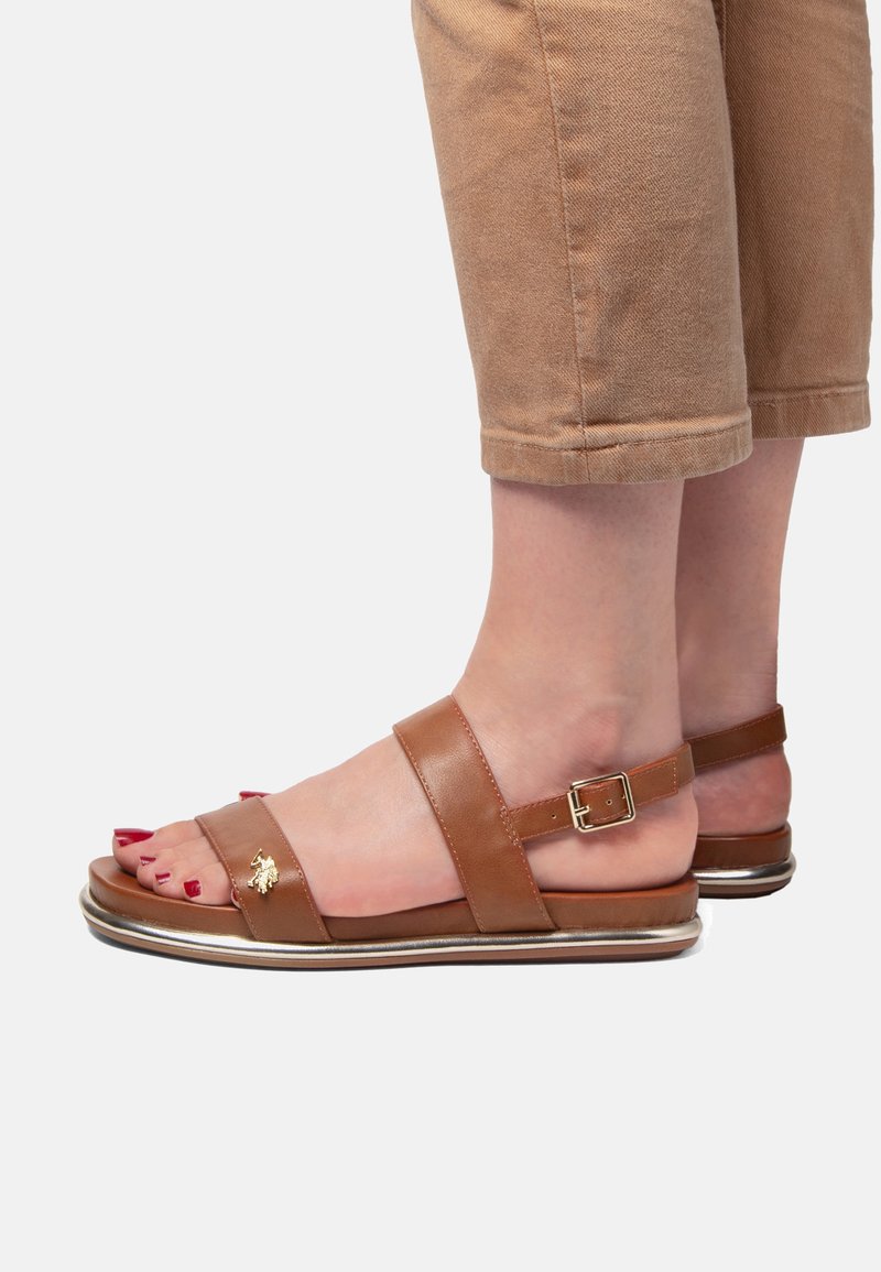 Hnědé kožené sandály se dvěma širokými pásky, s detailním sponkovým zapínáním a kovovým akcentem na podrážce. Odkryté prsty s červeným lakem na nehty.