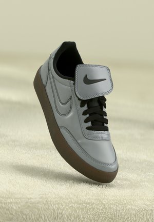 Šedé a černé tenisky Nike s hnědou podrážkou, černými tkaničkami a logem Nike na boku a jazyku, umístěné nad měkkým texturovaným povrchem.