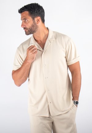 Chemise beige à manches courtes avec un motif texturé et un col décontracté. Associée à un pantalon assorti, offrant une coupe décontractée.